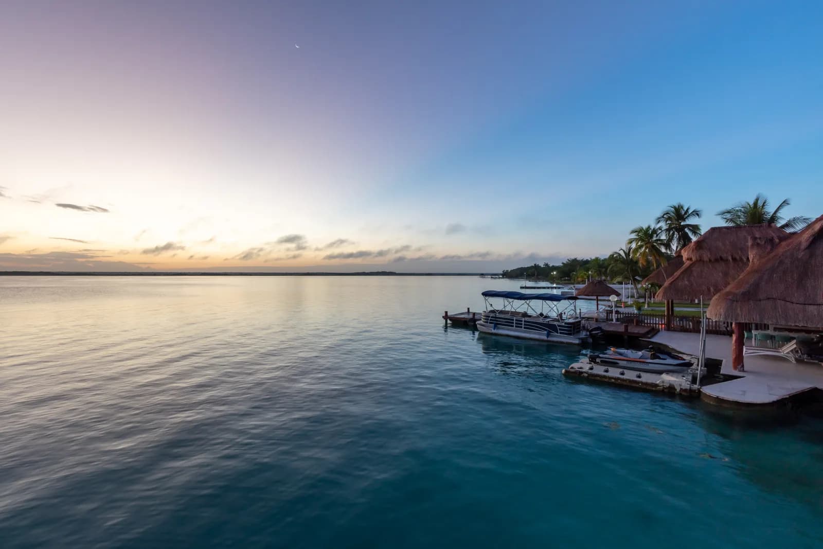 A pontoon tour boat moored at a palapa-roofed hotel dock on the Bacalar lagoon at dawn, Quintana Roo, Mexico