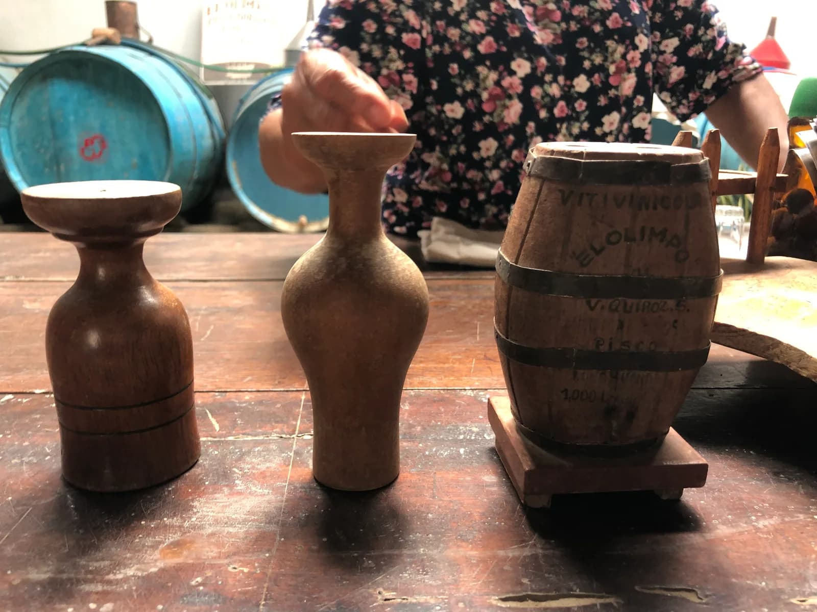 Wooden pisco measures and a small oak barrel on a worn table inside a family bodega in Lunahuaná, blue fermentation drums behind