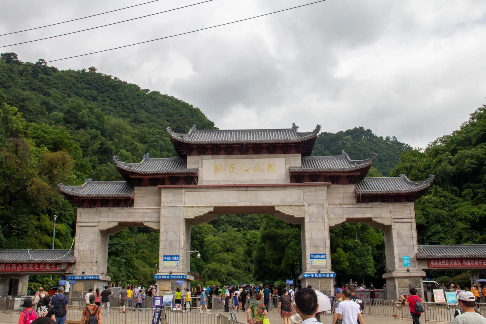 The stone and tiled-roof South Gate archway of Qianling Park in Guiyang with visitors queuing at the entrance lanes below forested hills