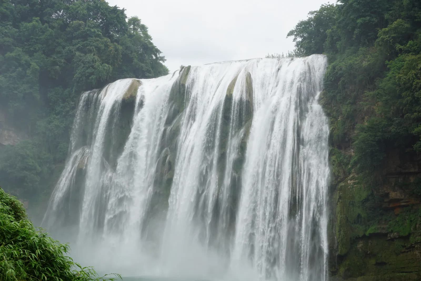 Huangguoshu Waterfall pouring over a wide limestone cliff into a turquoise pool surrounded by green karst hills